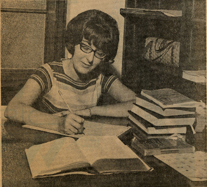 Photograph clipped from a newspaper showing Sally Priesand writing at a desk with books surrounding her.  