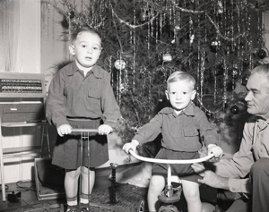 Two young boys on tricycles with a man holding one up on the right. A Christmas tree with tinsel is in the background.  