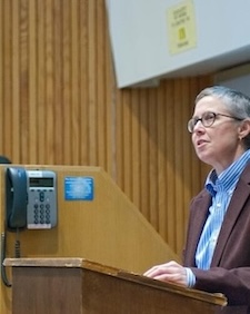 Emilye Crosby standing in front of a room, behind a podium, speaking.