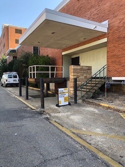 Photo taken on the street with a the library on the right side. The building has a loading area with steps and black metal posts around the loading area. Beside the steps is a shade the reads Cook Library Curbside Pickup. Further down the street is a white van.