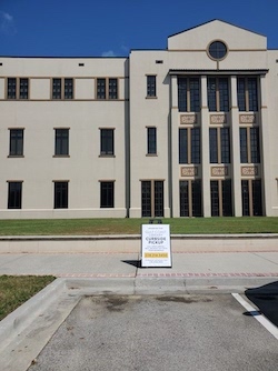 Photo has a large three floor off white building with three floors and windows. In front of the building is a side walk in a parking space. In front of the parking space is a sign that reads Curbside Pickup. On the left side of the parking space is a curb.