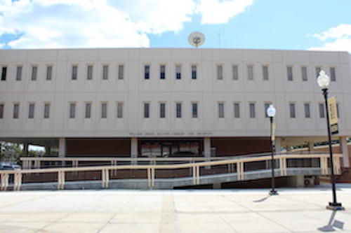 Photo is of the front of a building that is white concrete on the top portion and a covered walk around on the first level. Leading up to the first floor is a ramp that goes along in front of the building with concrete and iron railings.