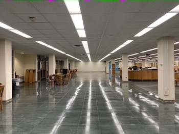 photo is a large room with green floors. On the left side of the photo are wooden tables with chairs next to a row of white columns. To the right side of the photo is another row of white columns with a large wooden desk in the center.