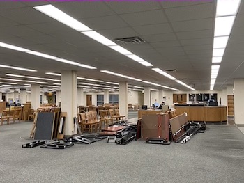 photo is a large room and in the center of the photo are stacks of tables that have been disassembled. Beside the stacks of table tops is a row of wooden chairs, surrounded by white columns. Beyond the tables and chairs is a large wooden desk.