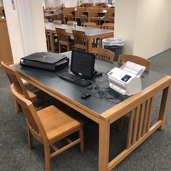 photo is of a wooden table with a green top and four chairs around the table. On top of the table is a black scanner, desktop computer with screen and keyboard, and to the right is a white printer. Beyond the table is a column and rows of tables and chairs.