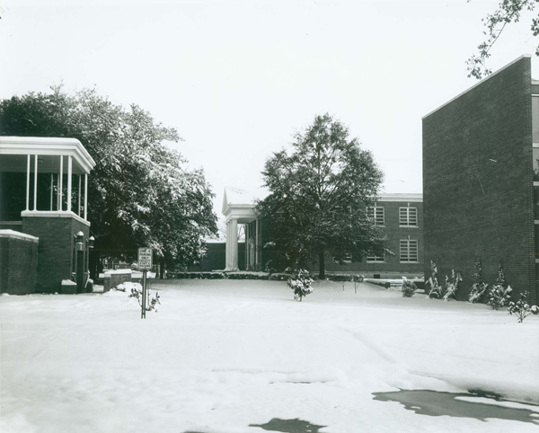 This photograph shows a vacant lot covered in snow on the campus of the University of Southern Mississippi. To the left of the lot is a brick porch that it is covered. To the right of the lot may be McLemore Hall on the Southern Miss campus. In the rear of the picture may be Kennard Washington Hall, which is a brick building with large white columns in front. In the foreground of the photograph is a parking sign reading “Zone 4 Parking Only. Faculty, Staff, and Visitors.”