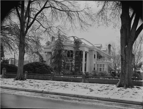 A large mansion sits off the road covered in snow. A large front yard featuring trees and bushes is also covered with snow. A concrete fence surrounds the property and sits approximately 10 feet from the road, which is in the foreground. There is some evidence of snow on the road. The mansion has three stories, appears to be brick, and has large white pillars in the front. There are numerous balconies off the building and a sun room is on the right side of the house. Two large trees stand in the foreground of the picture and are situated between the road and the fence.
