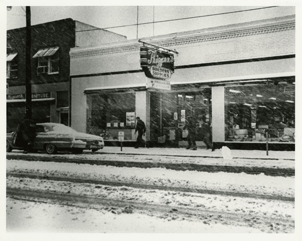 This photograph shows the store fronts of Thigpen’s Builder Supplies and Thigpen’s Furniture Store covered in snow. Only a half of the front of the furniture store shows in the photograph. It is a brick building with awnings over the windows. There are air conditioning units in the upstairs units. In front of the building is a circa mid-1960s car with someone getting in or out of the car. The building supply store is white with black accents and two large windows looking into the store. There are 2 people walking in front of the store fighting the snow storm. Slush is on the street in front of the store with evidence where vehicles have driven on the road to make gullies in the snow.