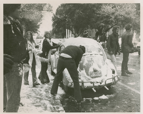 Two students are scraping snow off a VW bug. There is a guy in the rear of the car that has Alabama plates. A female student with a winter coat, bell bottoms and a knit hat is wiping down the driver’s side of the vehicle. Another woman is smiling to the left of the car while a male student, who is covered in snow, stairs pensively at something outside the picture. To the right of the car are seven students talking, laughing, and wondering around looking at the snow. There are also other cars in the photograph and College Hall, a brick classroom building with large white pillars, looms in the background.