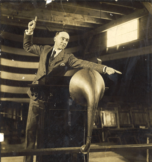 This photograph shows Howard S. Williams on an old, wooden stage pointing to the first or second row on his left. His right hand is pointing to the sky. In front of him is a podium with an early microphone devise that looks like a horn. Behind him is a section of a large American flag that only shows the lines of the flag. There are approximately eight rows of wooden folding chairs behind him with maybe 10 chairs per row. Behind him to the left is a window that is closer to the roof. Williams is wearing a tweed-looking suit with a vest, white button down shirt, and a polka dot tie. He has a pocket square in his suit pocket on the left.
