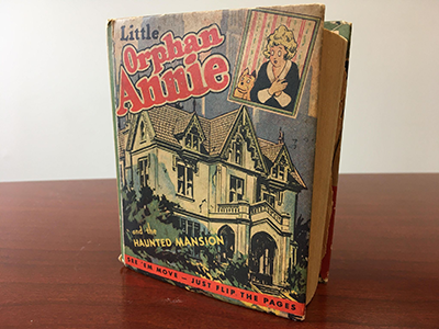 Book cover with title Little Orphan Annie and the Haunted Mansion, text in black and red. Text at the bottom of the cover says See ‘em move – just flip the page. Features a run-down house, and Annie and a dog look down at the house from the top right corner.