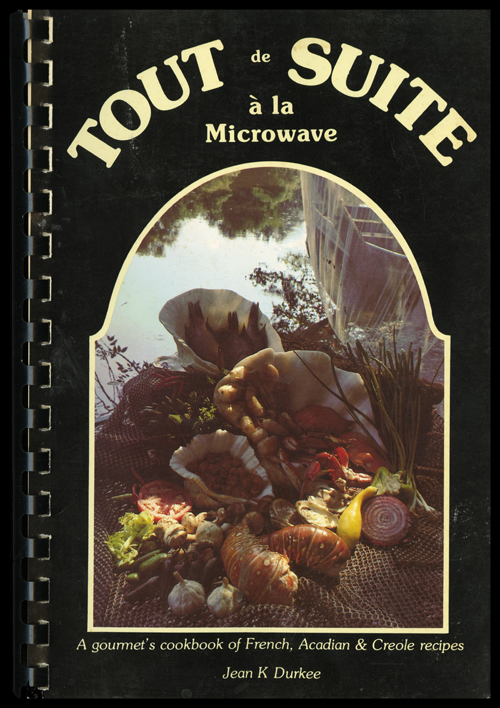 Cover of the cookbook Tout de Suite a la Microwave: A Gourmet’s Cookbook of French, Acadian & Creole Recipes by Jean K. Durkee. The cover is black with the title surrounding a cornucopia of food by a lake. 