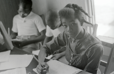 Female student in a Freedom School in Hattiesburg, Mississippi, looking at the camera with a pencil in her hand. Two boys in the back are writing in notebooks. 