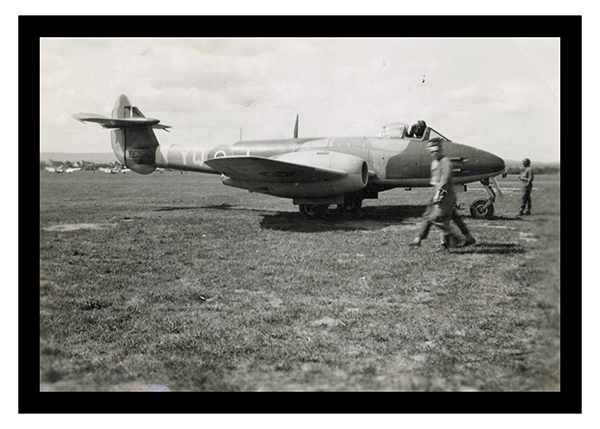 Black and white photograph of an airplane in a field. Two men are walking in the foreground with other soldiers surveying the airplane. 