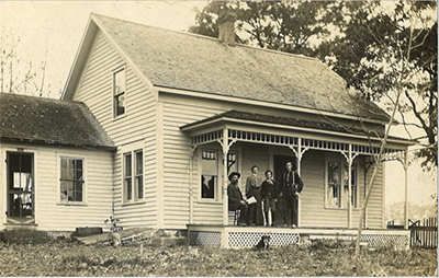 A black and white postcard depicting a family standing in front of an old house. There are three men and a woman standing on the porch.