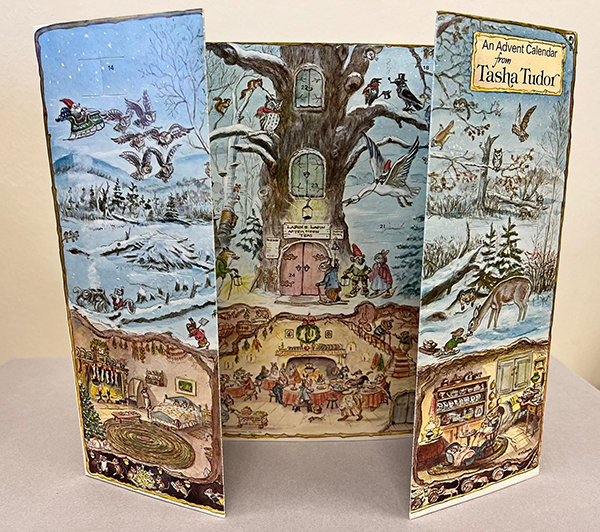 Unfolded, 5-panel, cardboard advent calendar with open-out, numbered cardboard windows. Text box in top right corner provides the title, An Advent Calendar from Tasha Tudor, in black text. Calendar features a winter woodland scene with various animals preparing food, walking or flying in the woods, and sleeping in beds. Central panel shows a sign above a tree (the tree has door in it) that says Lapin & Lapin Afternoon Teas, with a sign to the left labeled Menu and a sign to the right, above a bell, that says Please Ring. 
