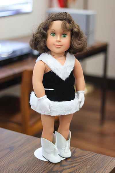 Two images of an 18-inch doll. The doll has green eyes, short brown hair with bangs on each side, and painted on eyelashes. She is wearing a black dance uniform with white fur trim, white gloves, and white boots with tassels on the front. The first image shows the entire doll, which is being held up by a white metal doll stand. The second image is a close-up, showing the doll from the torso up.