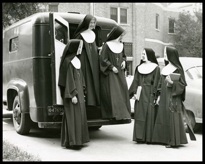 Three nuns are standing around the back of a paddy wagon while two nuns exit the vehicle. The license plate is from Michigan and appears to be in front of a building that may be a religious structure. 