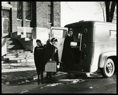 One nun is entering the rear of a paddy wagon while three other nuns make their way to enter. They are carrying religious books and other items to deliver to children. 