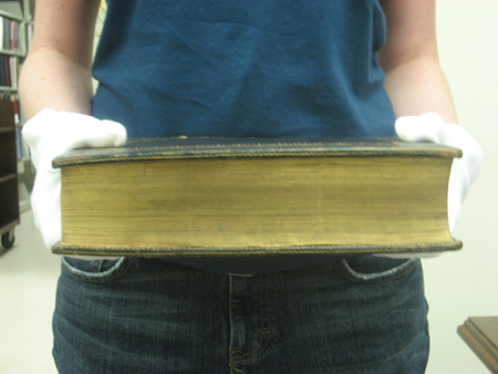 Book held by a person wearing white gloves showing the fore-edge of the book with the gold gilding.  