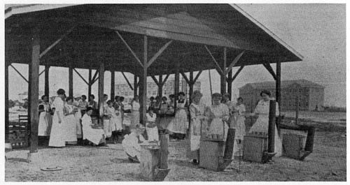 Members of the Tomato Club at Mississippi Normal College under a roof canning tomatoes. Early university buildings are in the background.  