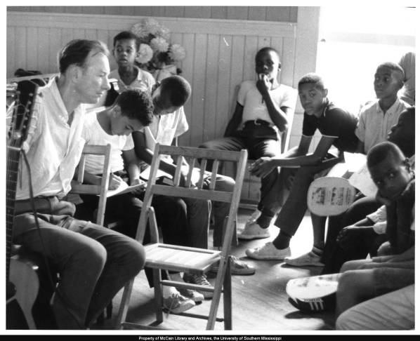 Folk singer Pete Seeger sits with his guitar in folding chairs with eight children surrounding him.  