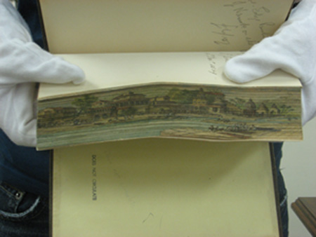 White-gloved hands fanning the fore-edge of a book showing the hidden painting. 