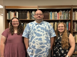 Three students, one male in the center, and two females standing in front of a tall shelf of books.