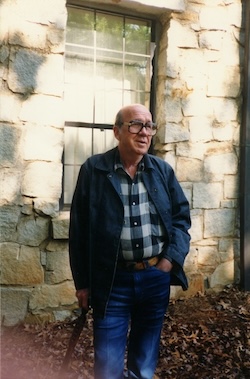 Photograph of Will Campbell, standing with hand in pocket, outside in front of a stone building, wearing a blue jacket, plaid shirt, and jeans.