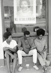 Black and white photograph of three black young adults, two female, with a male in the middle, sitting on a front step in front of a storefront, reading a notebook.