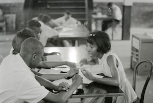 Black and white photograph of four black kids, one female on the right side of the table and three male on the left, talking and writing on paper.