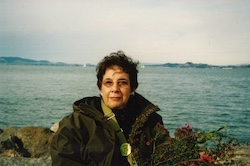 Sheila Michaels outdoors, in a coat, sitting in front of a lake.