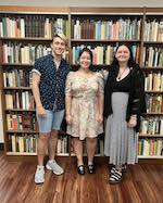 Three students, one male on the left, and two females standing in front of a tall shelf of books.