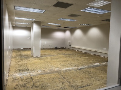 image is a photo of a room that has had the flooring removed. The walls are bare and a gray/off white color. A column stands in the middle of the room. Along the right side wall, wiring is exposed, and ceiling tiles have been removed, for construction purposes.