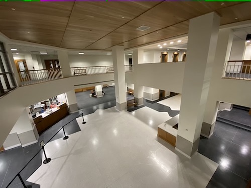 Photo is an overhead photo from the second floor, looking to the first floor. Floor on the lower level is white with black trim around the outer edges. Large white columns are around the edges of the room, with a railing on the second floor balconies. On the left side of the photo is the circulation desk, and to the right is the entrance to Starbucks.