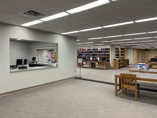 On the left side of the photo is an opening in a wall, a window, which is the service desk for research services. On the desk are two computers. On the right side of the photo are tables and chairs and beyond, that in the back, middle of the photo, are shelves with books.