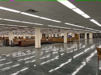photo is a large room with green floors and green carpet. At the front of the room is open floor, but at the back of the photos, beyond several white columns are tables, being taken down, lying on the floor, and a large wooden desk. To the right in the photo are a wall of windows.