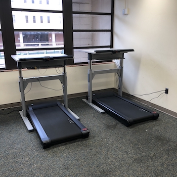 photo is of two treadmills with desktops to be used for walking and working. On the other side of the treadmills are horizontal windows. To the right is a white wall with an outlet where the treadmills are attached.
