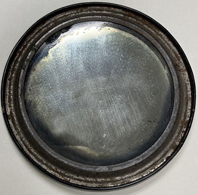 Back of the pocket mirror with a view of the mirror itself surrounded by a metal ring with indentation along the circular edge.